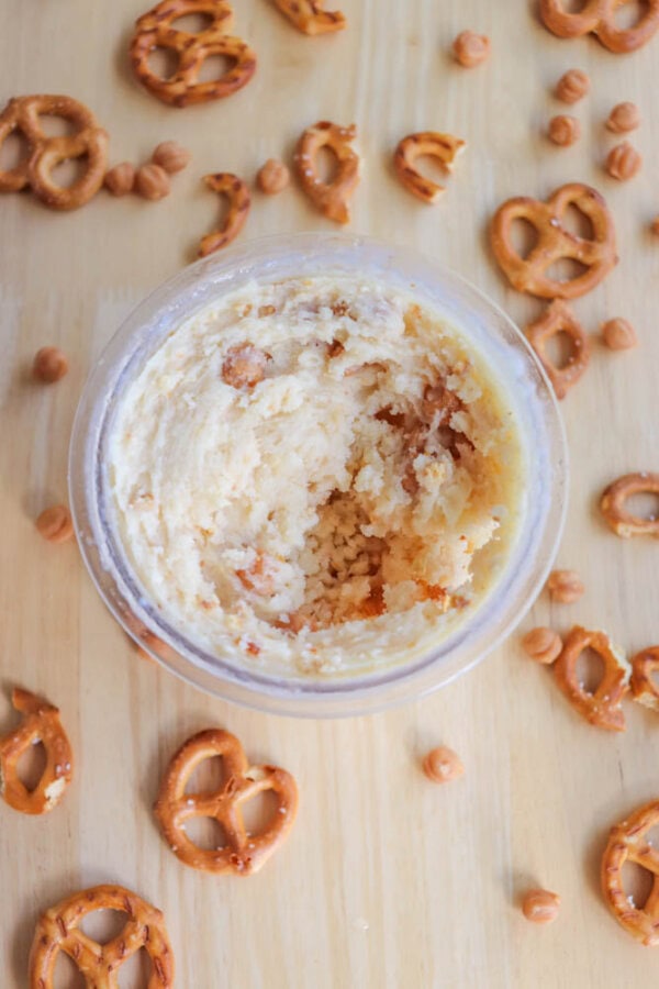 A jar of creamy ice cream with visible chunks sits on a wooden surface, surrounded by scattered pretzels and small caramel bits. The dip looks partially consumed, revealing its texture and ingredients.