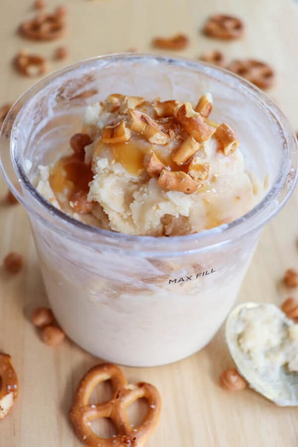 A cup of creamy vanilla ice cream topped with caramel drizzle and crushed pretzel pieces. A spoon with remnants of ice cream is on the wooden surface, surrounded by scattered whole pretzels and caramel bits.