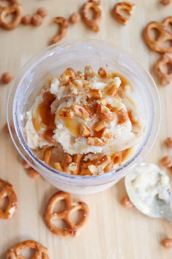 A cup of vanilla ice cream topped with caramel sauce and crushed pretzels, surrounded by whole pretzels and butterscotch chips on a wooden surface. A silver spoon with a bit of ice cream rests nearby.