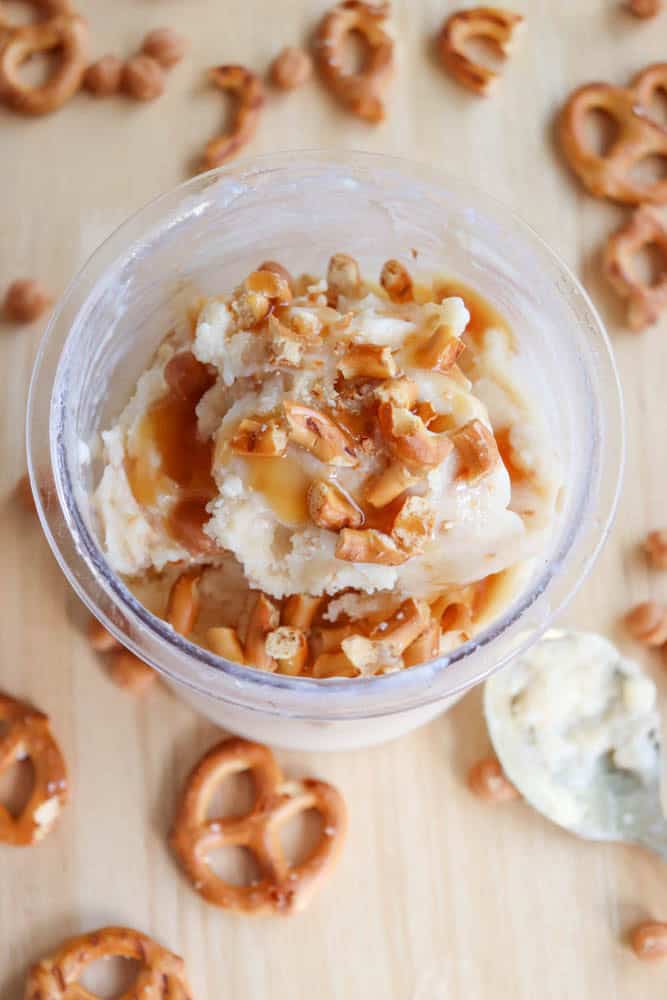 A cup of vanilla ice cream topped with caramel sauce and crushed pretzels, surrounded by whole pretzels and butterscotch chips on a wooden surface. A silver spoon with a bit of ice cream rests nearby.