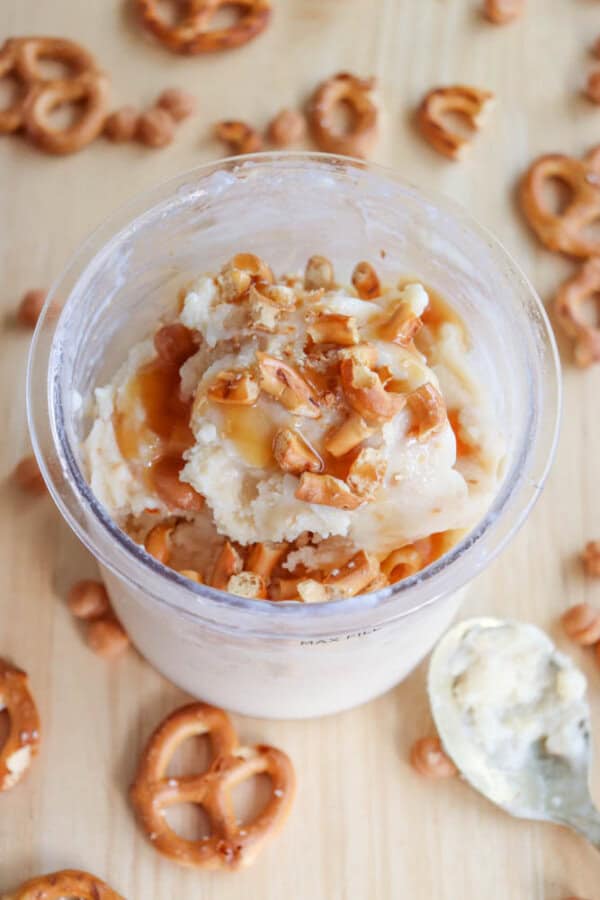 A clear cup filled with salted caramel ice cream topped with caramel sauce and crushed pretzels. Surrounding the cup are more mini pretzels and caramel chips scattered on a wooden surface. A spoon with ice cream rests beside the cup.
