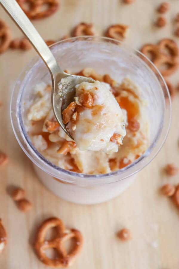 A spoonful of creamy dessert topped with caramel and small pretzel pieces, held over a cup filled with the same dessert. Scattered pretzels and caramel bits are visible on the wooden table surface.