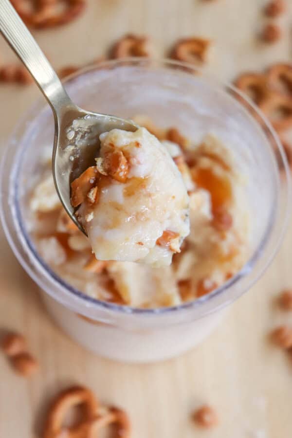 A spoon holds a portion of ice cream mixed with caramel swirls and pretzel pieces over a clear cup. Additional pretzel bits are scattered on the wooden surface.