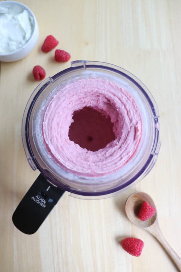 A ninja creami cup with creamy pink raspberry ice cream on a wooden surface. Nearby, a wooden spoon holds a raspberry. Additional raspberries and a bowl of white yogurt are scattered around.