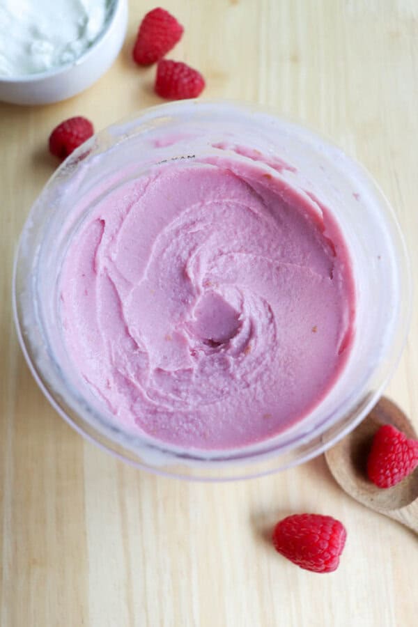 A bowl of smooth, pink frozen yogurt sits on a wooden surface. Raspberries are scattered nearby along with a wooden spoon holding one raspberry. A bowl of white cream is partially visible in the background.