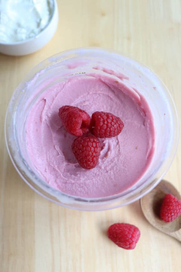 A bowl of pink raspberry frozen yogurt topped with fresh raspberries sits on a wooden surface. Nearby, a wooden spoon holds a couple of raspberries, and a small white bowl, partially in view, contains a white substance.
