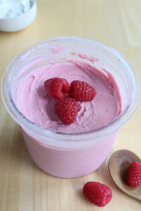 A container of pink raspberry yogurt with three raspberries on top sits on a wooden surface. Nearby, a wooden spoon holds two additional raspberries. A blurred white bowl is in the background.