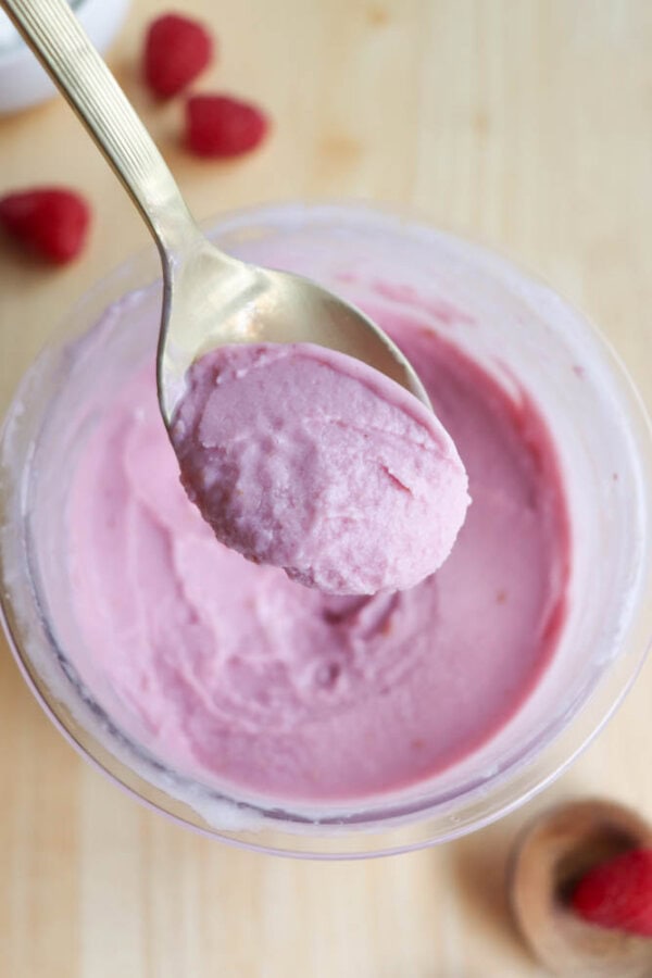 A spoon scooping pink, creamy raspberry frozen yogurt from a glass bowl. The yogurt has a smooth texture, and there are a few whole raspberries scattered on the light wooden surface near the bowl.