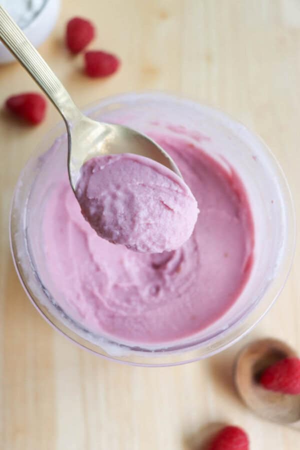 A spoon holds a scoop of pink raspberry frozen yogurt above a bowl filled with more of the yogurt. Several whole raspberries are scattered on the wooden table surface around the bowl.