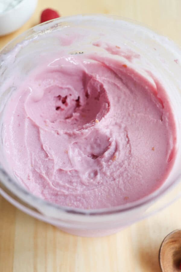 A close-up of a pint filled with creamy, light pink raspberry frozen yogurt. The ice cream has a smooth texture with a few small air pockets. The jar is placed on a light wooden surface.