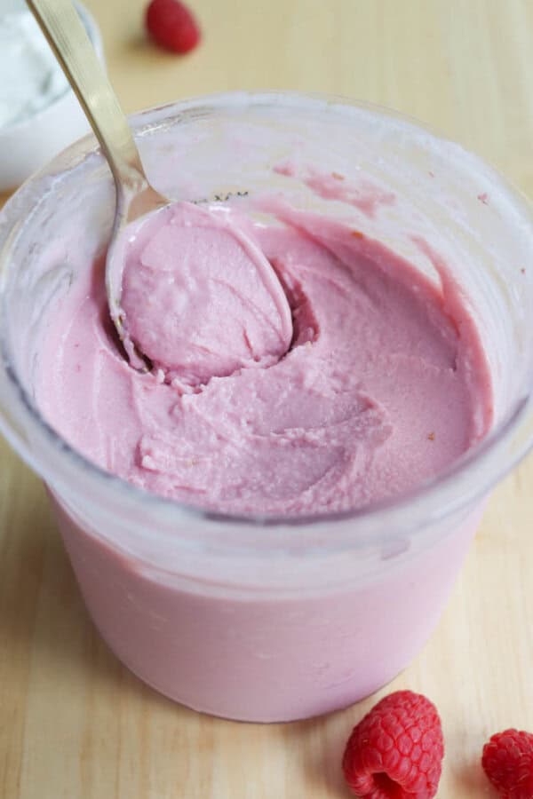A spoon rests inside a container of creamy pink yogurt on a light wooden surface. Fresh raspberries are scattered nearby, adding a touch of vibrant red. A bowl of white yogurt is partially visible in the background.