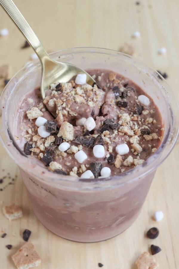 A clear cup filled with chocolate ice cream garnished with chocolate chips, mini marshmallows, and crumbled graham crackers. A gold spoon is dipped into the pudding. The cup is placed on a wooden surface with scattered toppings.