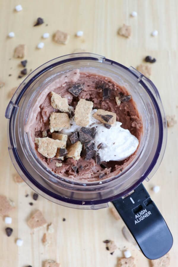 A ninja creami cup filled with chocolate ice cream topped with crushed graham crackers, chocolate chunks, and marshmallow fluff. More graham cracker pieces and chocolate chunks are scattered on the wooden surface around it.