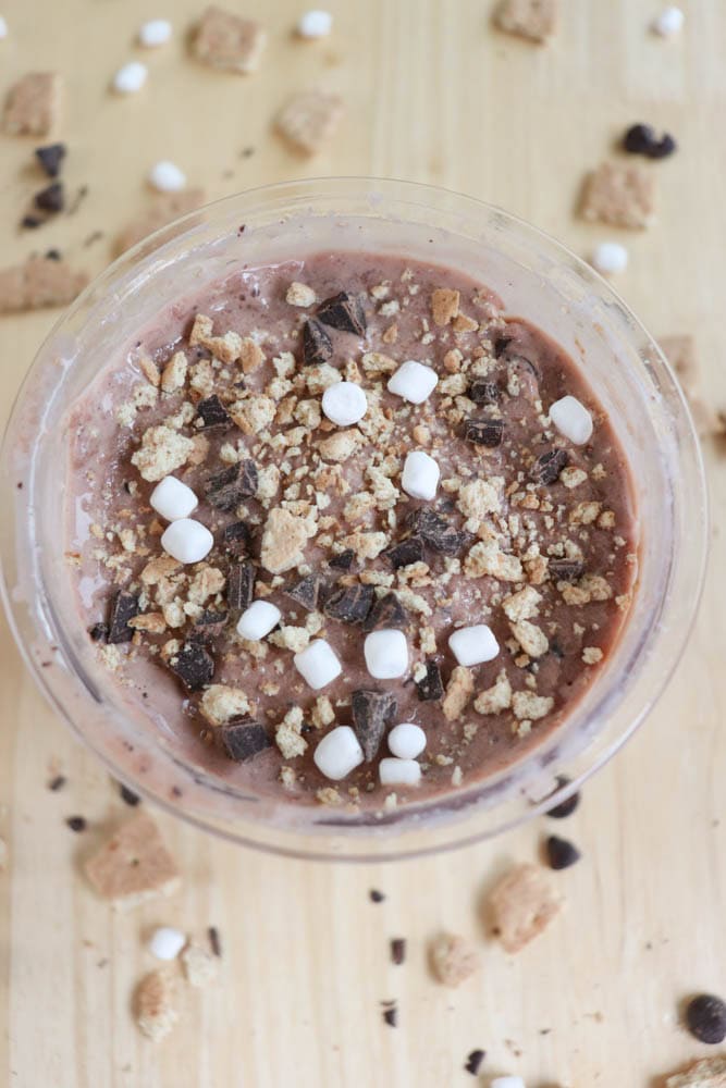 A bowl of s'mores ice cream with small marshmallows, chocolate chunks, and cookie crumbles. The surface is creamy and chocolatey, with additional toppings scattered on a light wooden table.