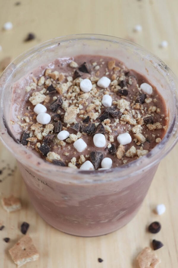 A clear cup filled with a chocolate dessert topped with mini marshmallows, chocolate chunks, and crumbled graham crackers, set on a wooden surface.