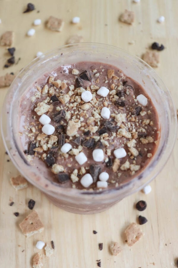 A bowl of chocolate dessert topped with mini marshmallows, chocolate chunks, and crushed pieces resembling graham crackers sits on a wooden surface, surrounded by scattered toppings.