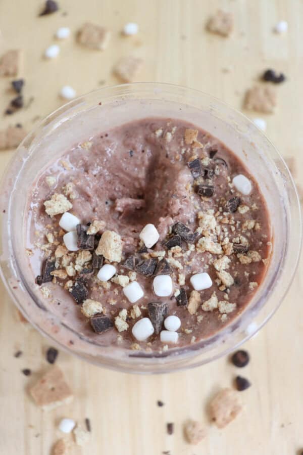 A bowl of chocolate smoothie topped with mini marshmallows, chocolate chips, and crumbled graham crackers on a light wooden surface. Crumbs and scattered toppings are visible around the bowl.