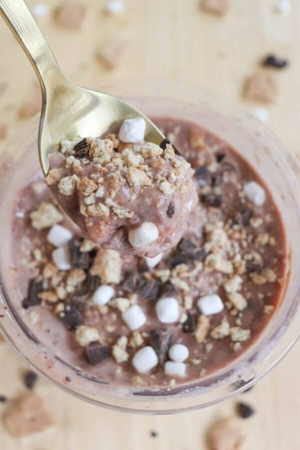 A pint filled with chocolate ice cream, topped with small marshmallows, crushed cookies, and chocolate chunks. A gold spoon rests in the pudding. The background features scattered cookie pieces.