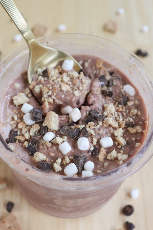 A creamy dessert topped with mini marshmallows, chocolate chunks, and crumbled cookies. A gold spoon is partially submerged in the dessert. The background features a few scattered marshmallows and chocolate pieces.