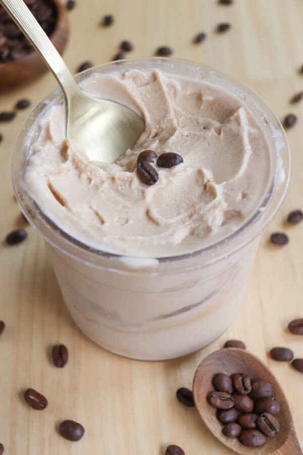 A clear cup filled with smooth coffee-flavored ice cream topped with a few coffee beans. A spoon is scooping some of the ice cream. Scattered coffee beans and a small wooden bowl with coffee beans are on the table.