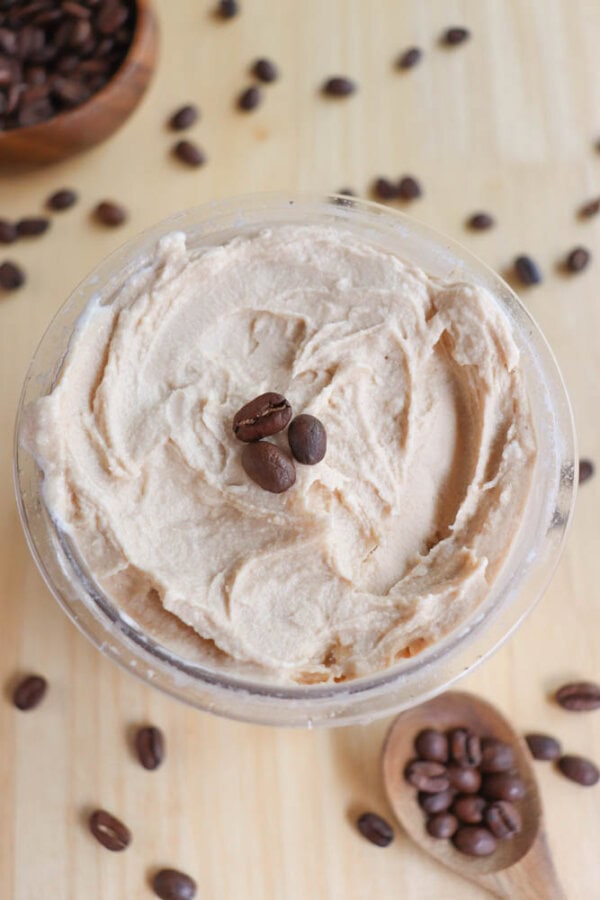 A bowl of creamy, tan coffee-flavored frosting topped with two coffee beans. Surrounding the bowl are scattered coffee beans and a wooden spoon filled with coffee beans, all on a light wooden surface.