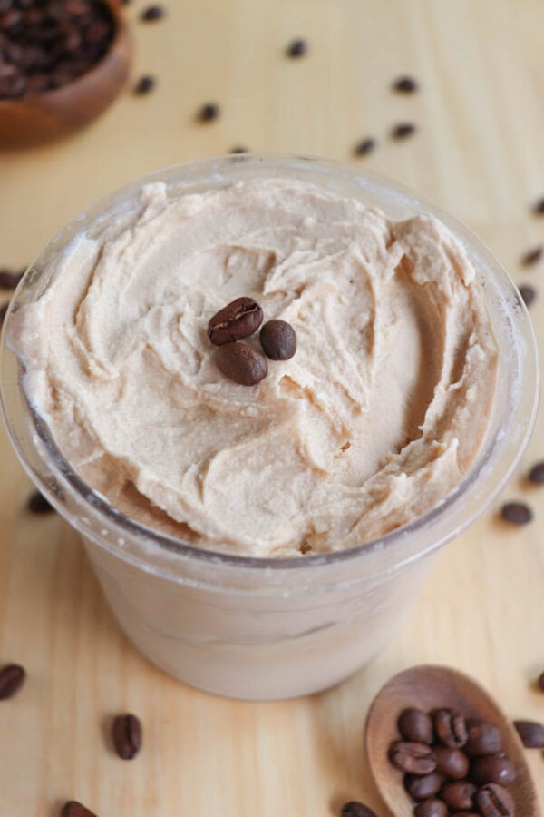 A cup of creamy coffee-flavored ice cream topped with two coffee beans is placed on a wooden surface. Scattered coffee beans surround the cup, adding a rustic touch.