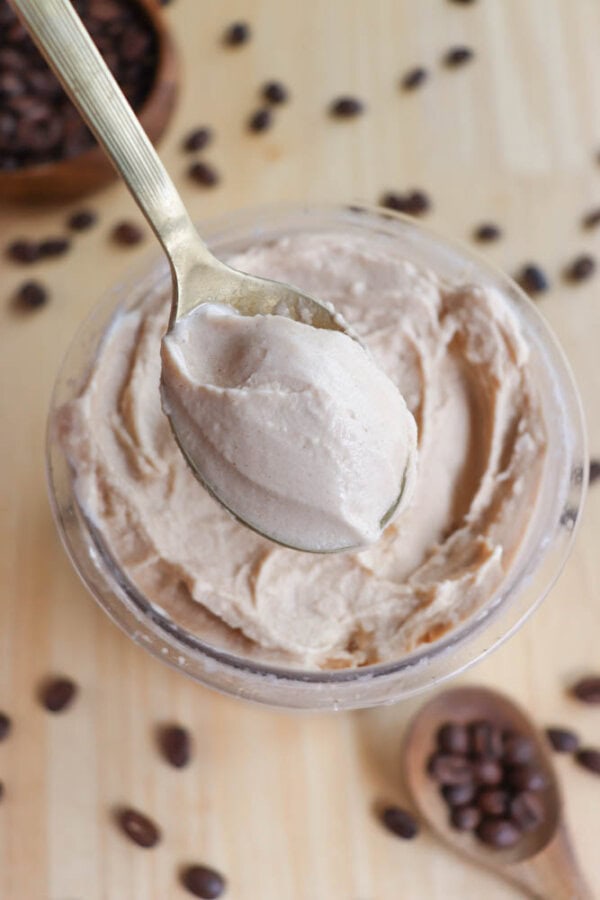 A spoonful of creamy coffee-flavored mousse is held above a glass container filled with the same mousse. Coffee beans are scattered on the light wooden surface around the container.