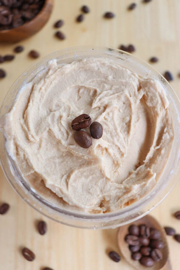 A bowl of light brown creamy spread, garnished with two coffee beans on top. Coffee beans are scattered around the wooden surface, with a wooden spoon holding more beans in the corner.