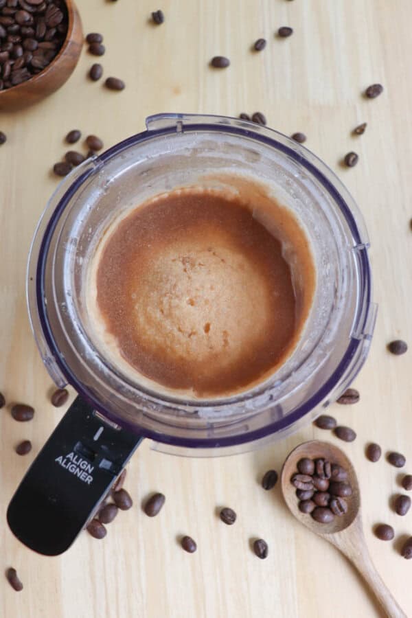 Aerial view of a blender containing a light brown mixture on a wooden surface. Coffee beans are scattered around, with some in a wooden spoon. A small wooden bowl filled with coffee beans is partially visible at the top left.