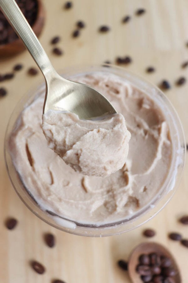A spoon holding a scoop of creamy coffee-flavored mousse is placed over a bowl filled with more of the same mousse. Coffee beans are sprinkled around the wooden surface, adding to the coffee theme.