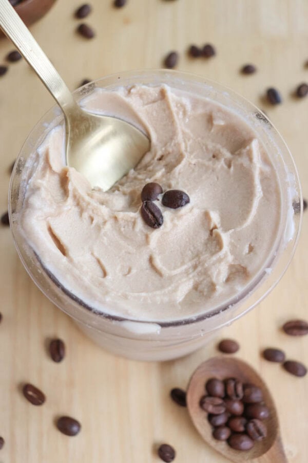 A container of creamy coffee-flavored ice cream with a gold spoon. Coffee beans are scattered around the container and placed on top of the ice cream. The background is a light wood surface with additional coffee beans.