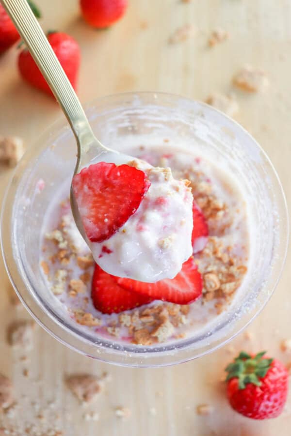 A spoon holding a strawberry slice with ice cream and graham cracker crumbs hovers over a pint. Inside, more strawberries, ice cream, and granola are visible. Whole strawberries and granola pieces are scattered on the light wooden surface like a raspberry cheesecake surprise.