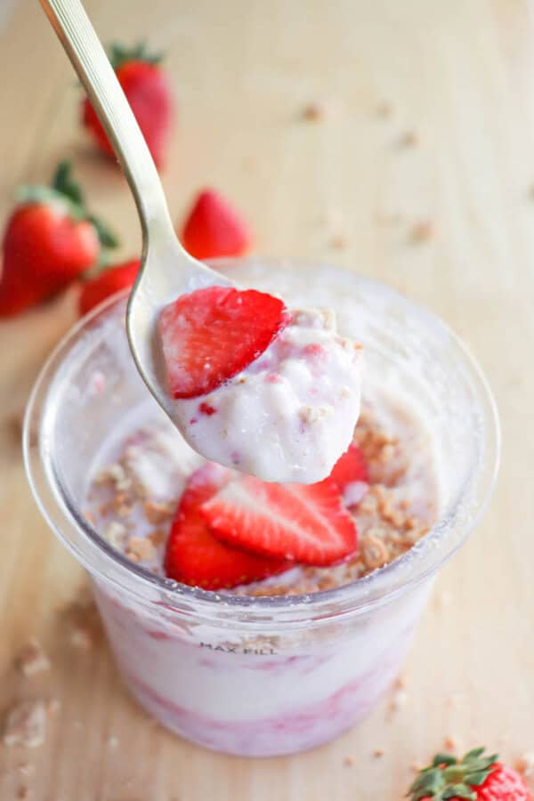 A spoonful of ice cream with sliced strawberries and graham cracker crumbs is lifted above a cup with the same ingredients, reminiscent of a refreshing raspberry cheesecake delight. Fresh strawberries are scattered on a light wooden surface in the background.