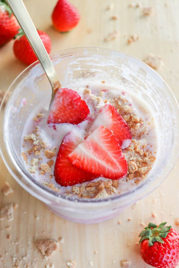 A glass cup filled with creamy ice cream is topped with graham cracker crumbs and sliced strawberries, resembling a delightful strawberry cheesecake. A spoon playfully peeks out, ready for a taste adventure. Whole strawberries are scattered around on the wooden surface.