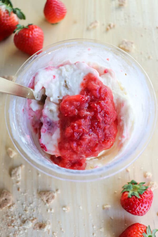 A bowl of creamy delight with a red strawberry topping is on a wooden surface. Strawberries and crumbly bits are scattered around the bowl, reminiscent of a cheesecake, while a spoon is dipped into the dessert invitingly.