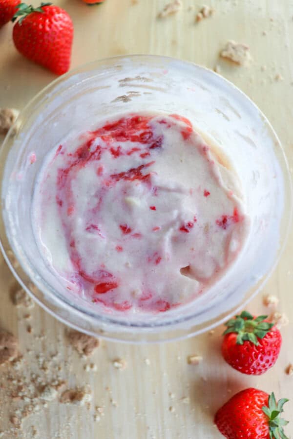 A bowl of strawberry ice cream mixture with visible strawberry pieces sits on a wooden surface. Fresh strawberries and scattered crumbs surround the bowl, reminiscent of a delightful raspberry cheesecake.