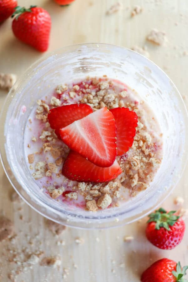 A bowl of creamy dessert reminiscent of a strawberry cheesecake, topped with crumbled cookies and sliced strawberries, rests on a wooden surface. Whole strawberries are artfully scattered around the bowl.