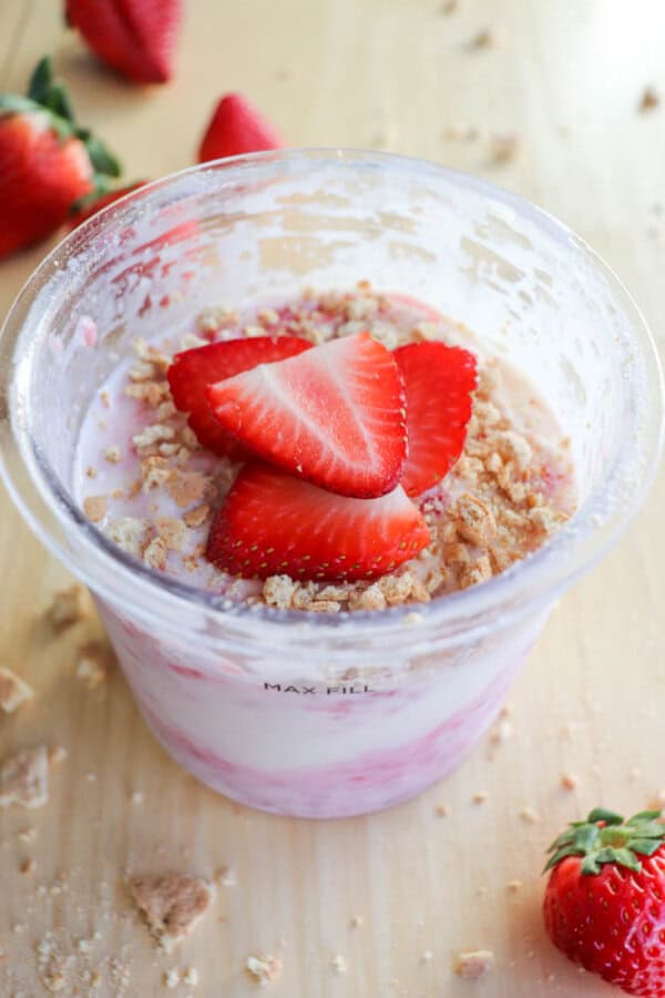 A plastic pint brimming with strawberry ice cream, elegantly topped with sliced strawberries and crumbled graham crackers. The scene captures a hint of raspberry cheesecake delight, all set on a wooden surface adorned with scattered whole strawberries.
