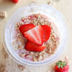 A top view of a pint filled with strawberry ice cream, crowned with sliced fresh strawberries and crumbled cookies. Whole strawberries are scattered on a light wooden surface, embodying the flair of a raspberry cheesecake in this delightful creation.