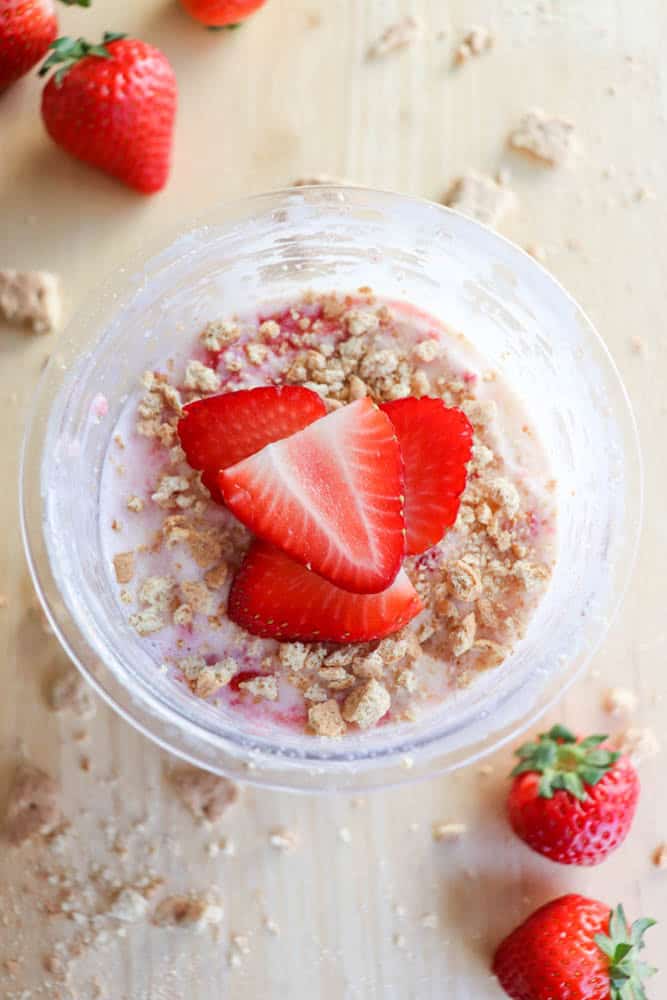 A top view of a pint filled with strawberry ice cream, crowned with sliced fresh strawberries and crumbled cookies. Whole strawberries are scattered on a light wooden surface, embodying the flair of a raspberry cheesecake in this delightful creation.