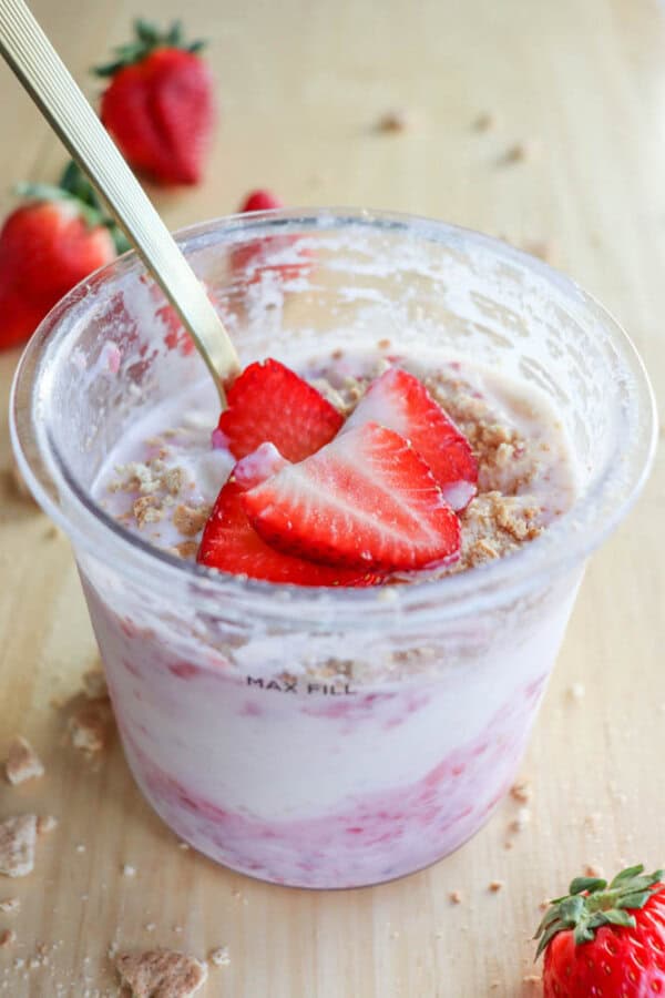 A cup of strawberry ice cream topped with fresh strawberry slices and crumbled biscuits resembles a raspberry cheesecake delight. A gold-colored spoon is dipped into the yogurt, as fresh strawberries are scattered on the wooden surface around the cup.