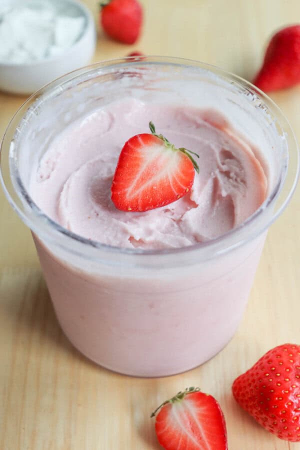 A clear plastic container filled with creamy strawberry yogurt is topped with a fresh strawberry half. Additional whole strawberries are scattered around on a light wooden surface. A small bowl of white yogurt is blurred in the background.