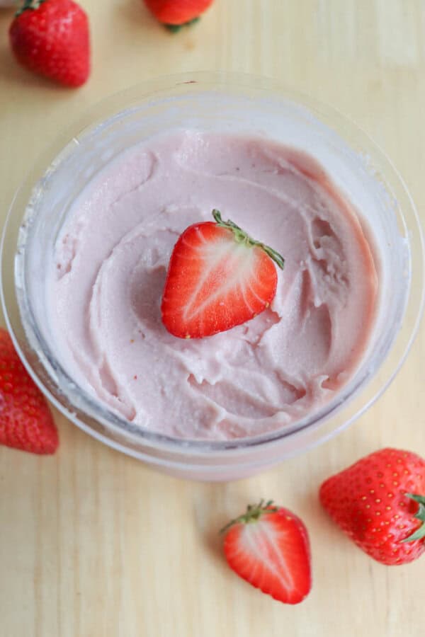 A clear bowl of creamy pink strawberry frozen yogurt is topped with a halved strawberry sits on a wooden surface. Fresh strawberries are scattered around the bowl.