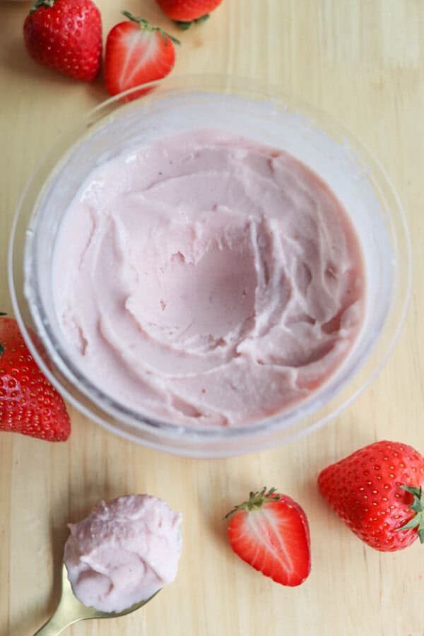 A bowl of creamy pink strawberry frozen yogurt on a wooden surface, surrounded by whole and halved fresh strawberries. A spoonful of the ice cream rests nearby.