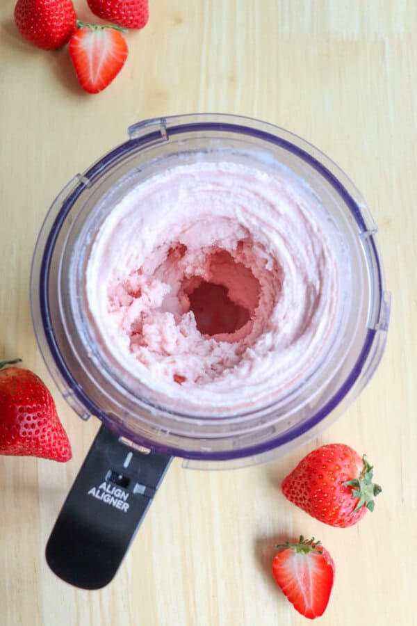 Top view of a food processor containing pink ice cream mix. Fresh strawberries are scattered around on a light wooden surface.