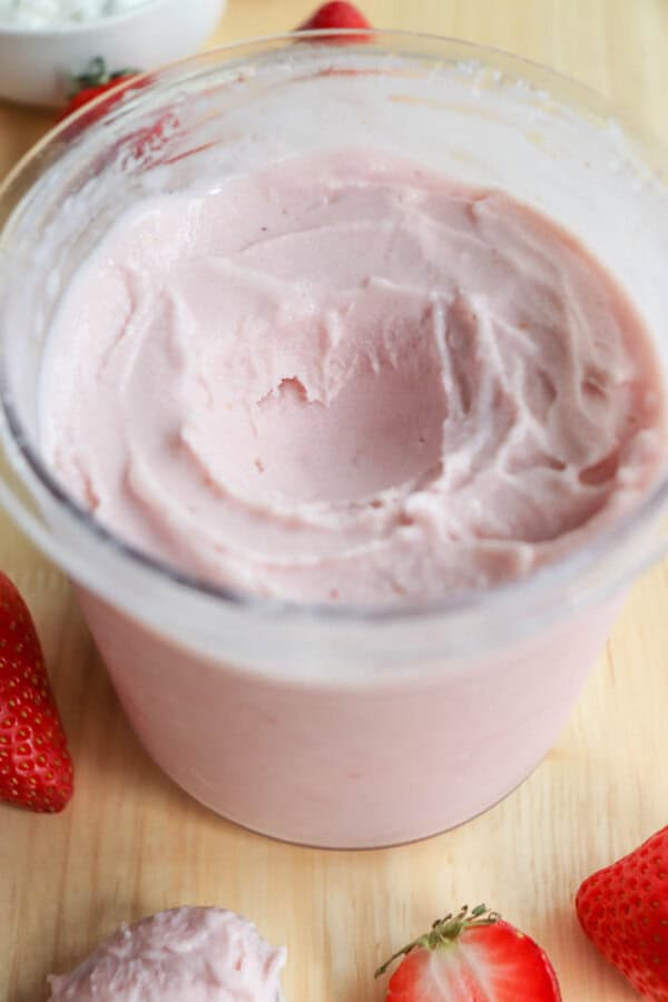 A container filled with creamy pink strawberry frozen yogurt on a wooden surface. Fresh whole strawberries and a halved strawberry are scattered around the container.