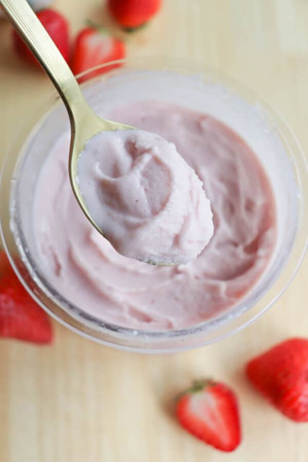 A spoon holding a scoop of pink strawberry frozen yogurt is positioned above a bowl filled with more of the yogurt. Fresh strawberries are scattered on the light wooden surface around the bowl.