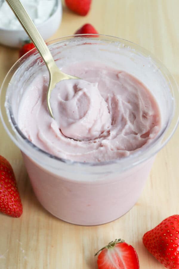A spoon rests inside a container of creamy frozen yogurt, surrounded by fresh strawberries on a wooden surface.