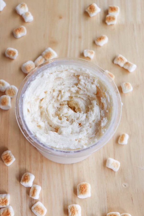 A glass jar filled with creamy marshmallow butter sits on a wooden surface. Small toasted marshmallows are scattered around the jar.