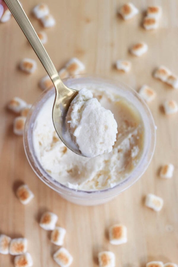 A spoon with a gold handle holds a scoop of creamy ice cream above a clear container. Small toasted marshmallow pieces are scattered on a wooden surface around the container.